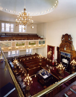 Interior view of the sanctuary from the women’s gallery. The dome in the ceiling, the huppa frame, the artwork on the gallery, the bimah, and the ark can be seen here. 